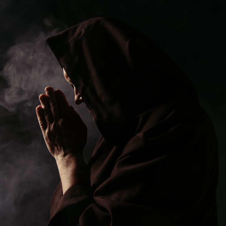 side view of priest with face obscured by hood praying on black with smoke
